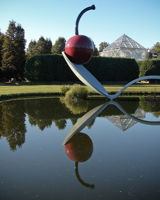 A picture of a very large sculpture of a bent spoon with a cherry in the dish of the spoon. All of this is over a lake at the Minnesota Sculpture Garden.