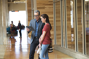 Older male college professor holding a Dell Venue 11 Pro 5000 Series (Model 5130 Midland) tablet computer and showing it to a young female college student, standing in the hallway of a campus building.