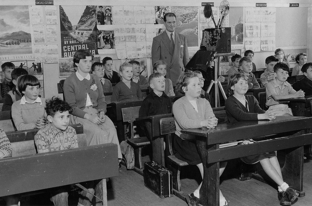 Students in a classroom from the mid -20th century. Teacher has a movie projector.