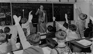 Old picture of a teacher in front of a class of young students. Many of them are raising their hands eager to answer a question.
