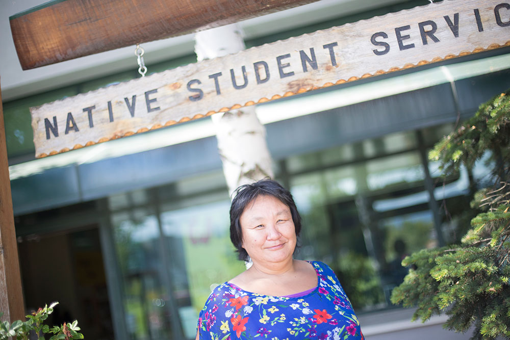 Eva Greg standing in front of a sign on intentionally weathered wood that reads "Native Student Services". She is a Native Alaskan with black hair and a few white specs in it.