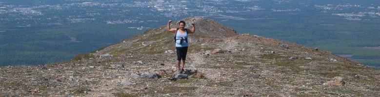 Eva raises her hands in triumph at the top of a peak with no trees. Beyond her is the vast vista of Alaska.