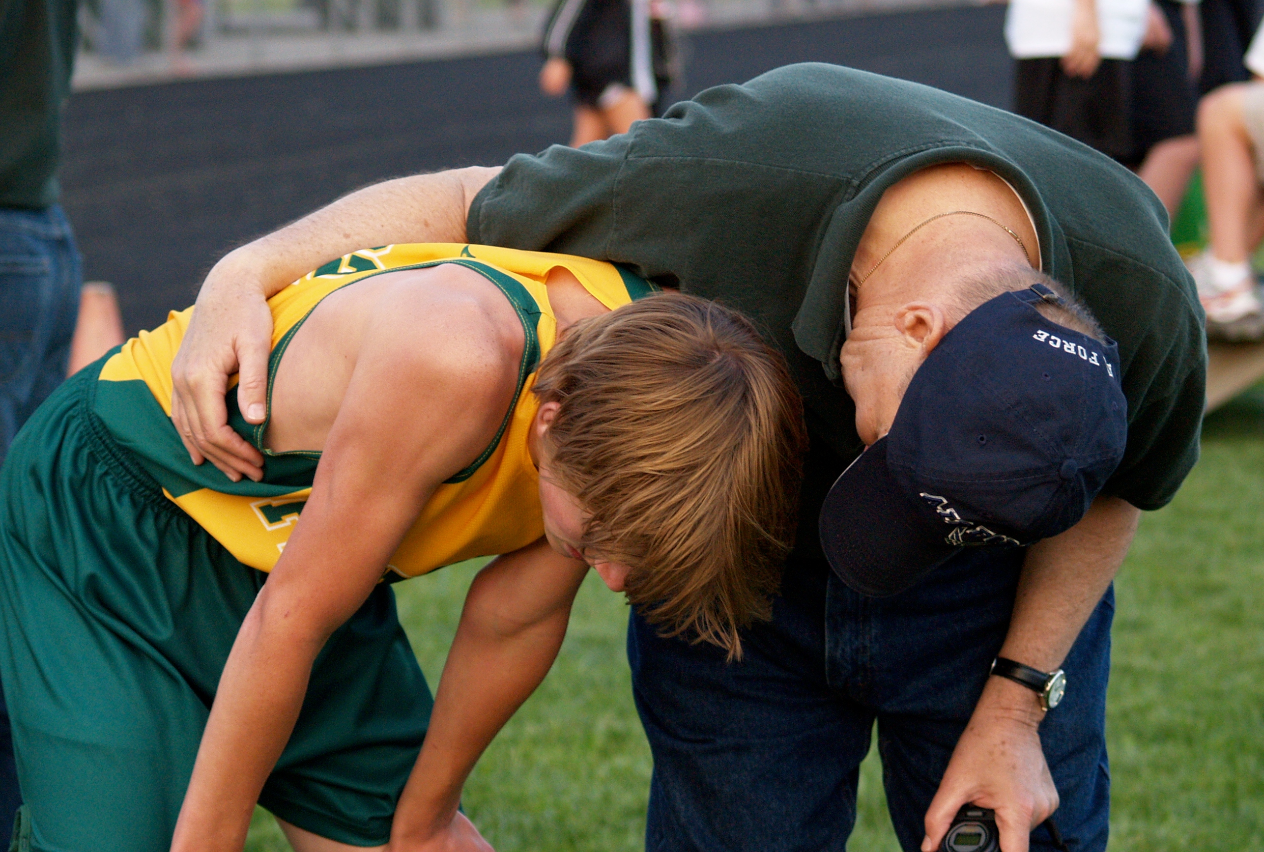 A track coach is leans down next to a young runner who, apparently just completed a race. The coach has his arm around the runner and appears to be consoling him.