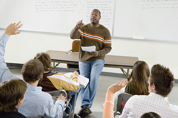 picture of a teacher in the front of a classroom of students seated in rows. The teacher is calling a student who is raising his hand.