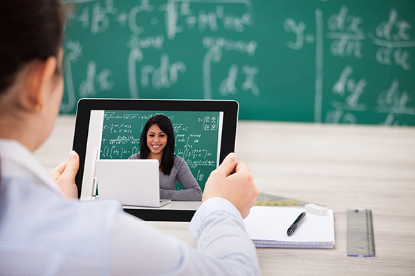 Picture of a faculty person looking at a tablet with a student on video on the tablet. The faculty person is facing a blackboard with mathematical formulas and the student has a similar blackboard with formulas behind her.