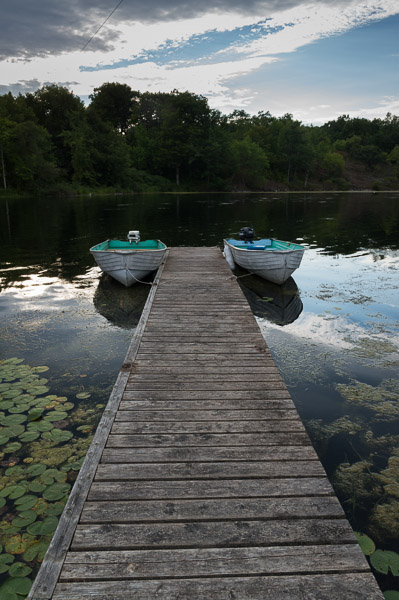 A photo of a lake with two vintage baots, tethered to a long, wooden plank. Behind the boats is lake water, and a bank of the lake with trees. 