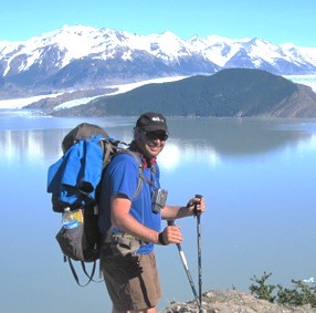 Author headshot. Doug stands in front of a mountain lake with snow covered mountains in the background.