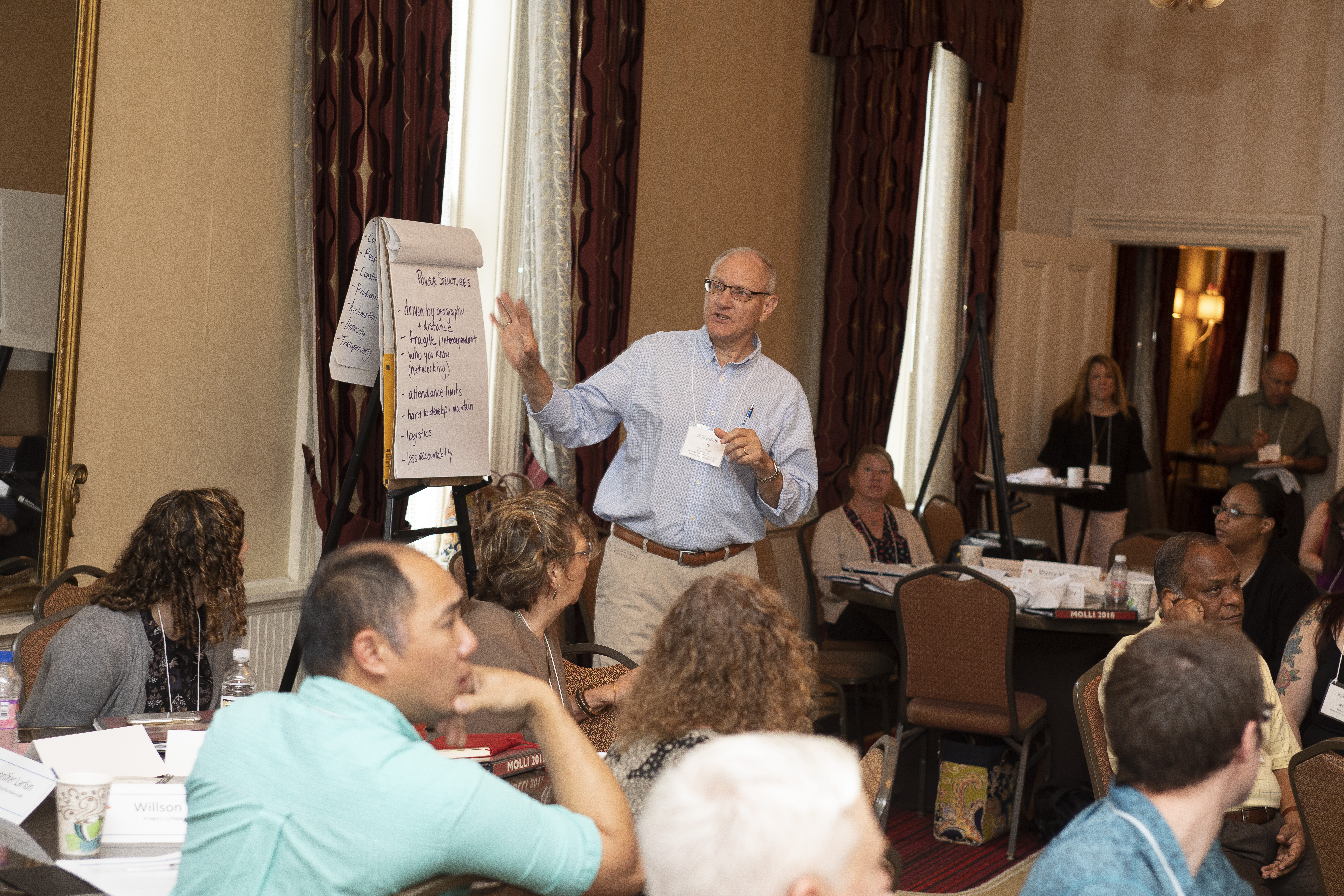 Participants in the 2018 institute presentation. Several individuals sit around round tables and one man presents from a paper flipchart with text written on it.