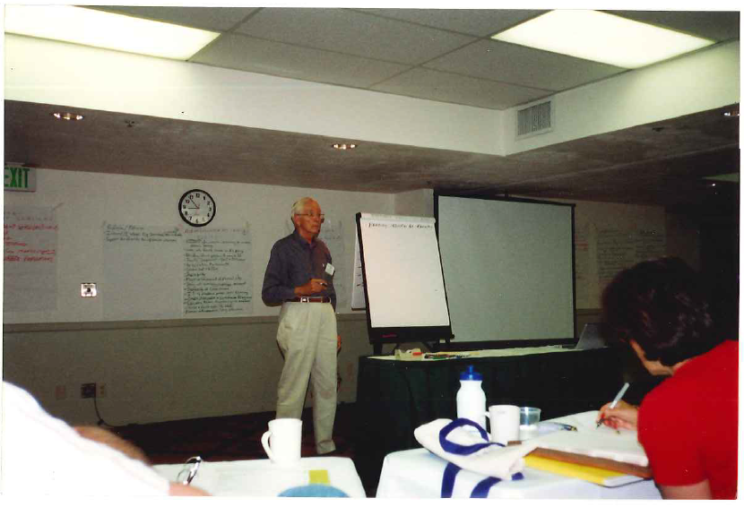 John Witherspoon, the first WCET Steering Committee chair, who was instrumental in setting some of the earlier foundations of WCET. He stands with a microphone in front of a classroom, next to a large notepad and a projector screen.