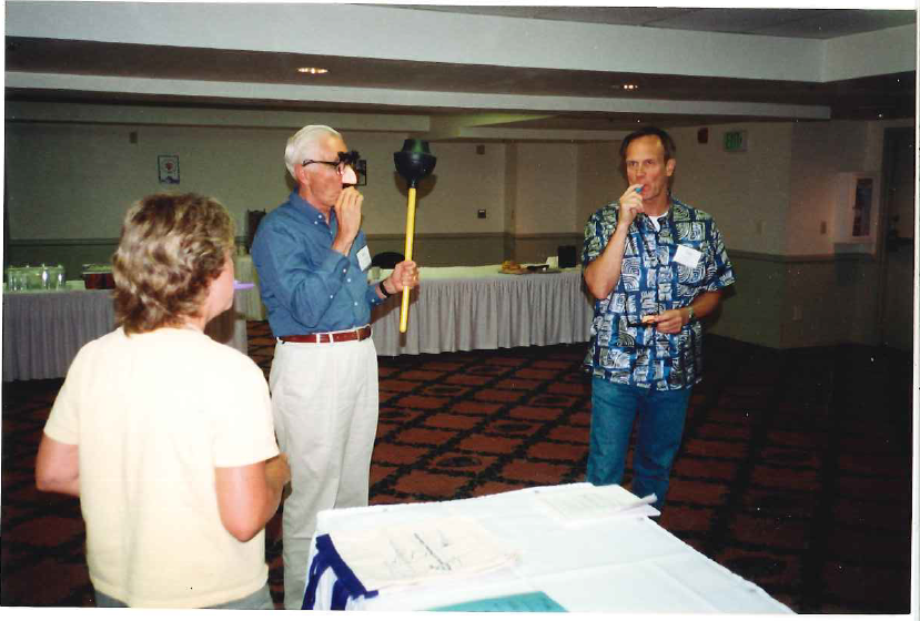 Darcy, John, and Ray practicing “pomp and circumstance” prior to the ceremony above. Three people stand in a room. One is wearing fake glasses, a fake nose, and a mustache and holding a plunger, another is blowing into a kazoo to make music.