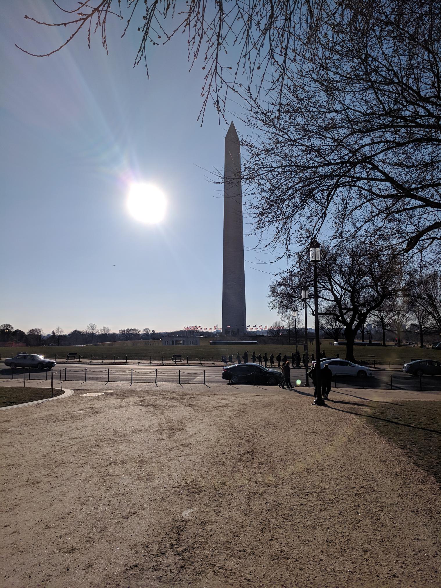 Picture of the Washington Monument from afar. The sun in shining behind the monument.
