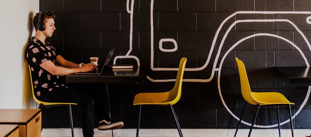 Picture of man sitting at a table and working on a laptop computer. He is wearing headphones and drinking coffee.