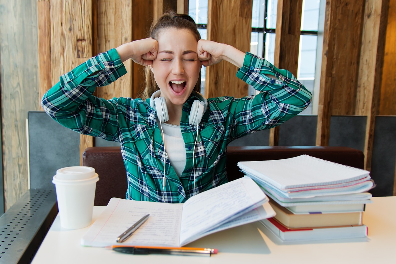 A young woman holds her head with her hands and screams. she is surrounded by notebooks as if studying.