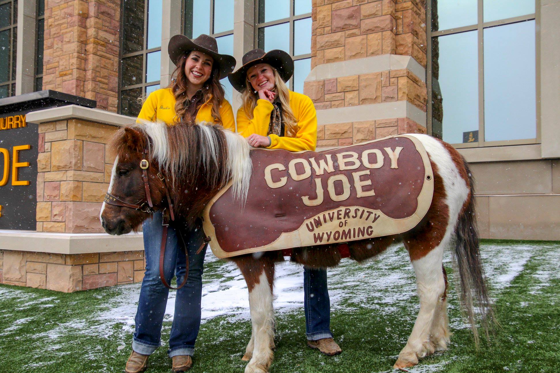 A pony with a cover that says Cowboy Joe University of Wyoming, posing with two women in cowboy hats