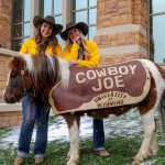 A pony with a cover that says Cowboy Joe University of Wyoming, posing with two women in cowboy hats