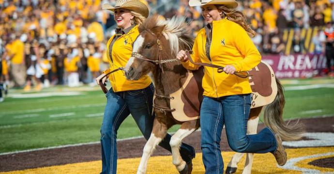 Two women in cowboy hats run with Cowboy Joe, the pony, on a football field 