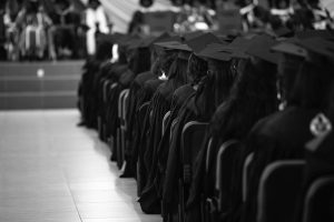black and white photo taken from behind several students in graduation caps and gowns sitting in chairs