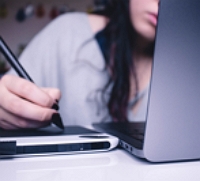 young person looks at a laptop and writes with a pen on table next to computer. 