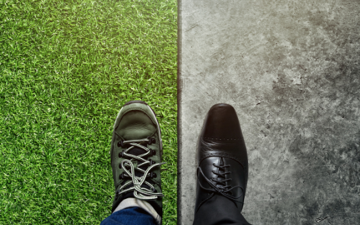 View of two feet standing with one foot in sneakers standing on grass, one foot in professional shoes on a sidewalk.