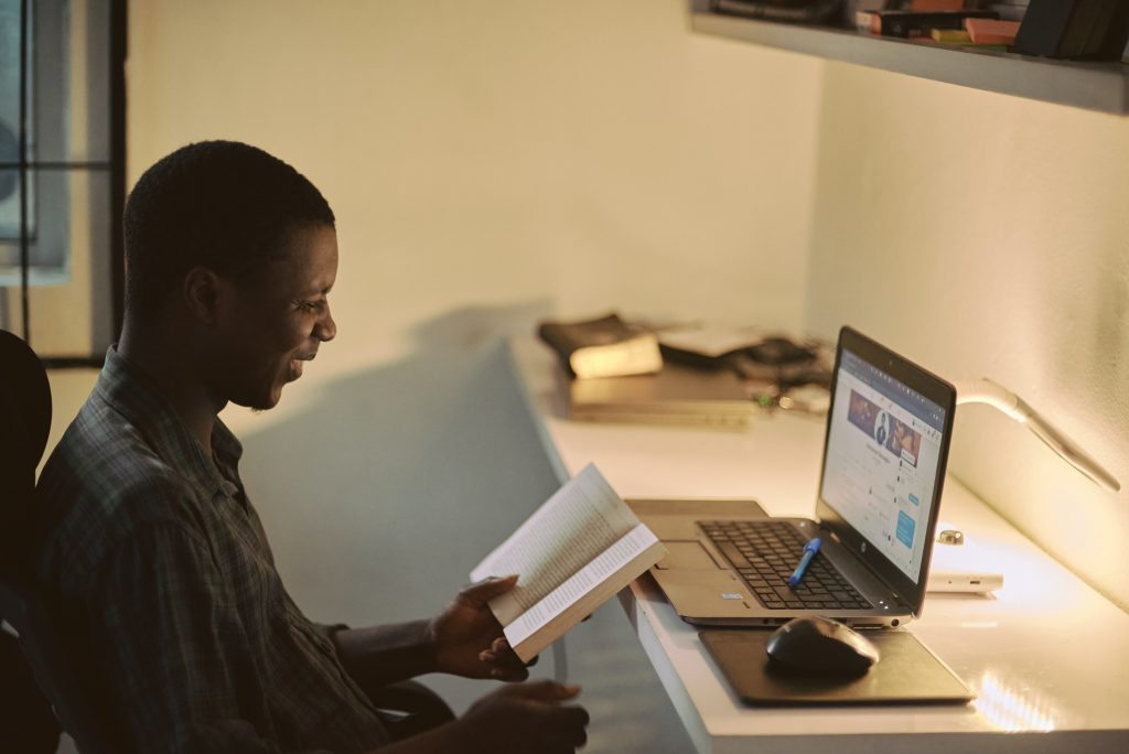 A younger man holding a book smiles at a laptop on a desk.