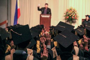 a speaker stands in front of a group of students wearing graduation caps and gowns. 