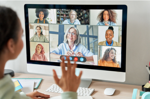 a student waves to a computer screen showing a video conference session with several young adults who may be fellow students and one individual who may be the instructor.