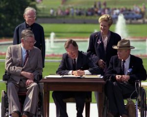 President George Bush sitting at a table on a lawn, surrounded by two individuals in wheelchairs and two others. Bush is signing something on the table.