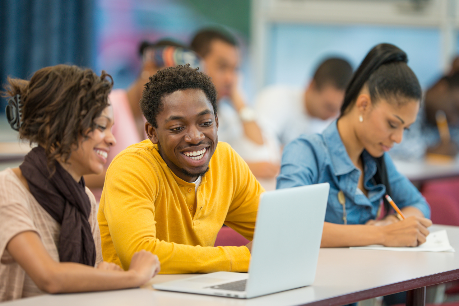 Students sit at desks in a classroom, two smiling at a laptop while another writes in a notebook.