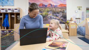 A smiling teacher and a young girl look at each other while sitting at a desk with a laptop in a brightly decorated early childhood classroom.